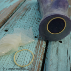 Photo shows a discarded condom with the base ring torn off laying next to a large purple silicone dildo that has been covered entirely in a condom, from tip to base