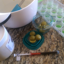 DIY setup for making my own vaginal THC suppositories like Foria Relief. Photo shows finished domed nuggets of pea-green cocoa butter, a mini ice cube mold, mixing bowl, jar of cocoa butter and syringe of medical THC oil.
