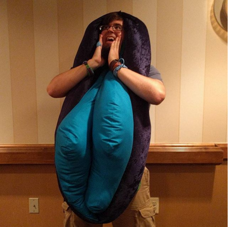 This is a photo of Kenton wearing a giant plushie vulva, and his head is where the clitoris would be. He's emulating Zach of Buzzfeed who wore a similar costume in a Buzzfeed video and adorably realized his head was the clitoris and was very excited. 